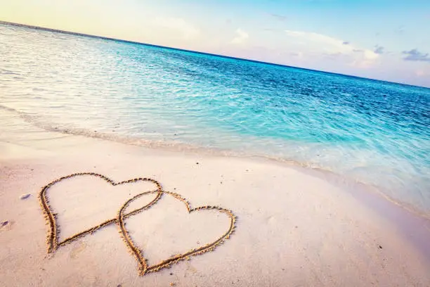 Photo of Two hearts drawn on sand of a tropical beach at sunset.