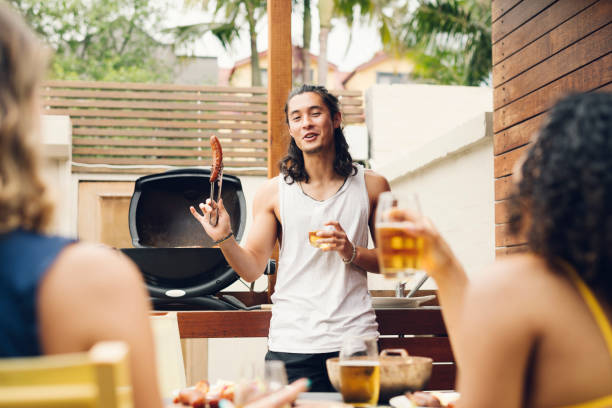 Young man holding up sausage in front of barbecue with friends on foreground Mixed race man in his 20s cooking food, smiling and talking, serving cooked sausage and drinking beer teasing photos stock pictures, royalty-free photos & images