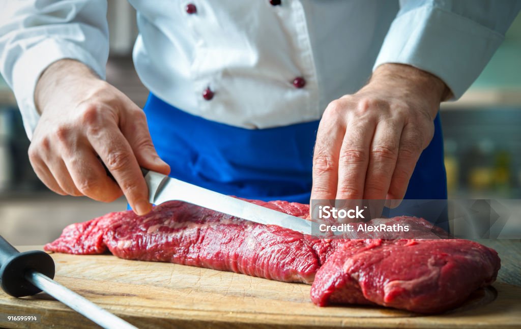 Hands of male chef in uniform cutting big piece of beef Hands of male chef in uniform cutting big piece of beef on board in restaurant kitchen. Man cuts meat with knife. Big piece of raw meat. Chef working in the kitchen Meat Stock Photo