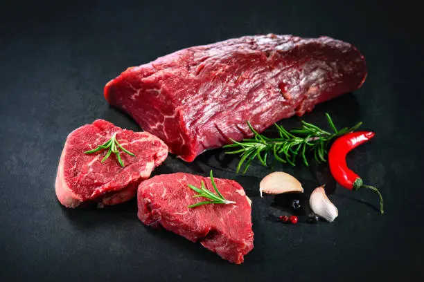 Photo of Whole piece of tenderloin with steaks and spices ready to cook on dark background