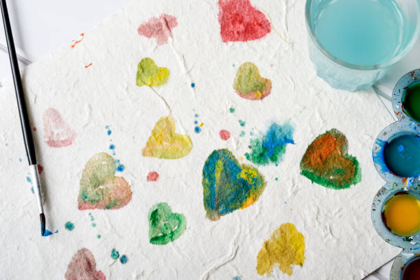 high angle view of a piece of paper with some hearts of different colors painted by myself with watercolor paint, next to a paintbrush and a palette with paint of different colors