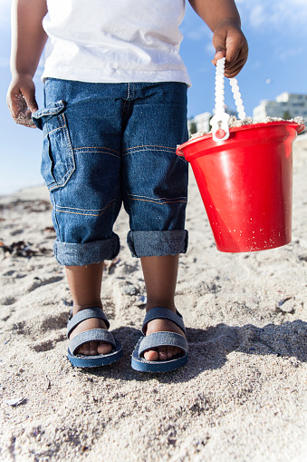 African child standing on the beach while he carries a full bucket of sand, preparing to build a sand castle.