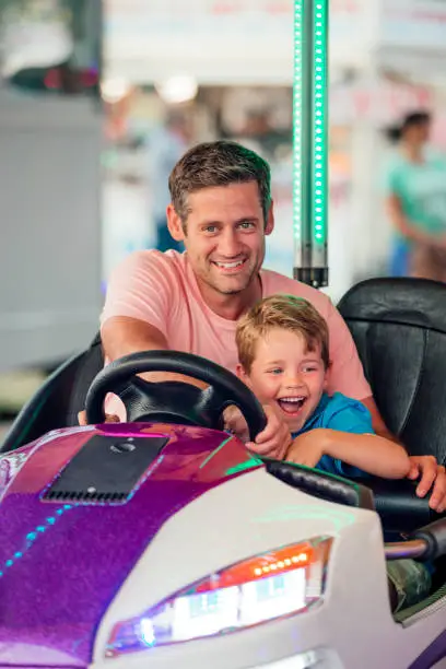 Mid Adult Man is behind the steering wheel of a dodgem car with a little boy at his side. They are at the fairground.
