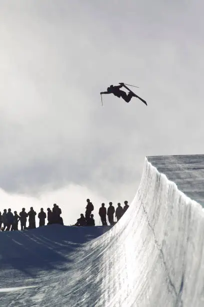A male alpine skier catches big air off a half pipe jump while other skiers look on from above.