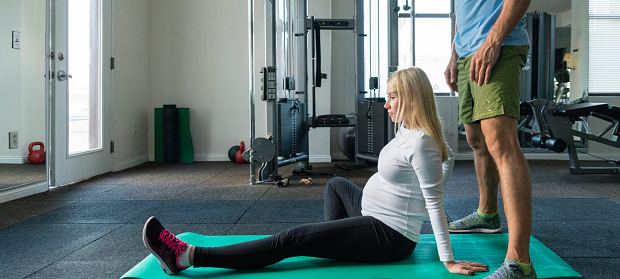 The athletic man, personal trainer, do fitness stretching relaxing neck and shoulders massage for a pretty pregnant woman in the gym.
