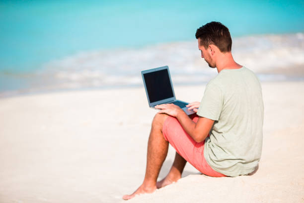 istock Young man sitting on sand with laptop on tropical caribbean beach. Man with computer and working on the beach 916660408