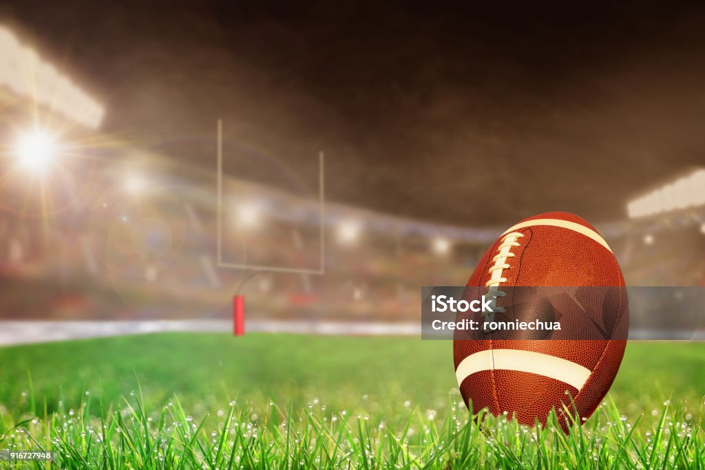 Outdoor Football Stadium With Ball on Grass and Copy Space American football on field grass in brightly lit outdoor stadium with focus on foreground and shallow depth of field on background. Deliberate lens flare and copy space. Fictitious football stadium created entirely in Photoshop. American Football - Sport Stock Photo