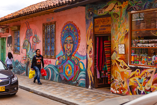 Bogota, Colombia - May 28, 2018: A local Colombian man walks through a colorful street in the historic La Candelaria District in the capital city of Bogota in the South American country of Colombia, quite unmindful of the brightly painted walls. Many of the street facing walls in the area are painted in the vibrant colours of Colombia, sometimes depicting Pre-Colombian legends or modern Street Art and Graffiti. The mural in the backgound, to the left was done by the popular Colombian Street Artist called Rodez. Some Graffiti touts have unfortunately defaced the mural by signing over it. Photo shot in the late afternoon sunlight in the Golden Hour; horizontal format. Camera: Canon EOS 5D MII. Lens: Canon EF 24-70mm F2.8L USM.
