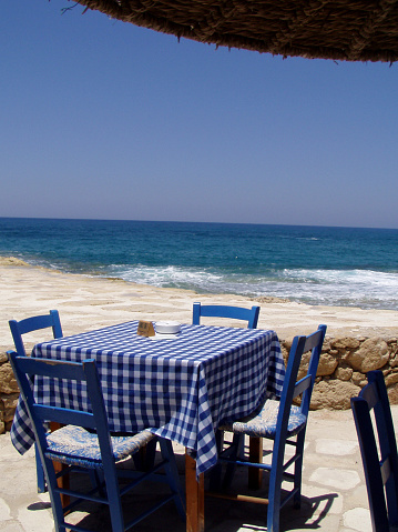Idyllic  view on Free table in the cafe and chairs  in colors of Greece - white and blue. People swimming in distance  and yachts (sailboats) anchored behind
