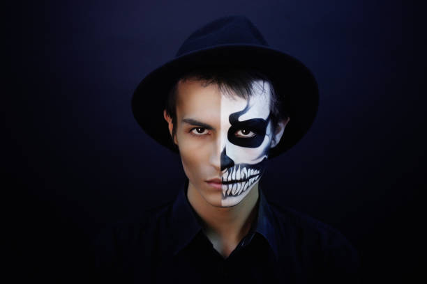 Creative man with make-up for the Halloween party. The original idea for Halloween black and white skull on his face, half of the image. Professional portrait near black wall, free place horizontal