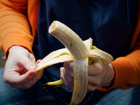 Person wearing jeans is sitting on the floor and eating banana