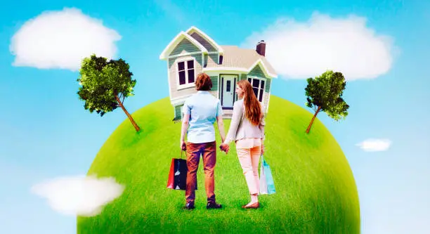 Man and woman are ready to buy a new home. The are holding shopping bags and looking at a house on green lawn. The couple is bonding and holding hands.