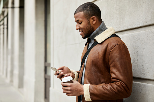 Smiling man with disposable coffee cup and mobile phone against wall. Young male tourist is wearing leather jacket. He is exploring city.