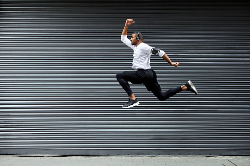 Full length of determined young man jumping against shutter. Side view of fit male jogger is exercising in mid-air. He is wearing sports clothing.