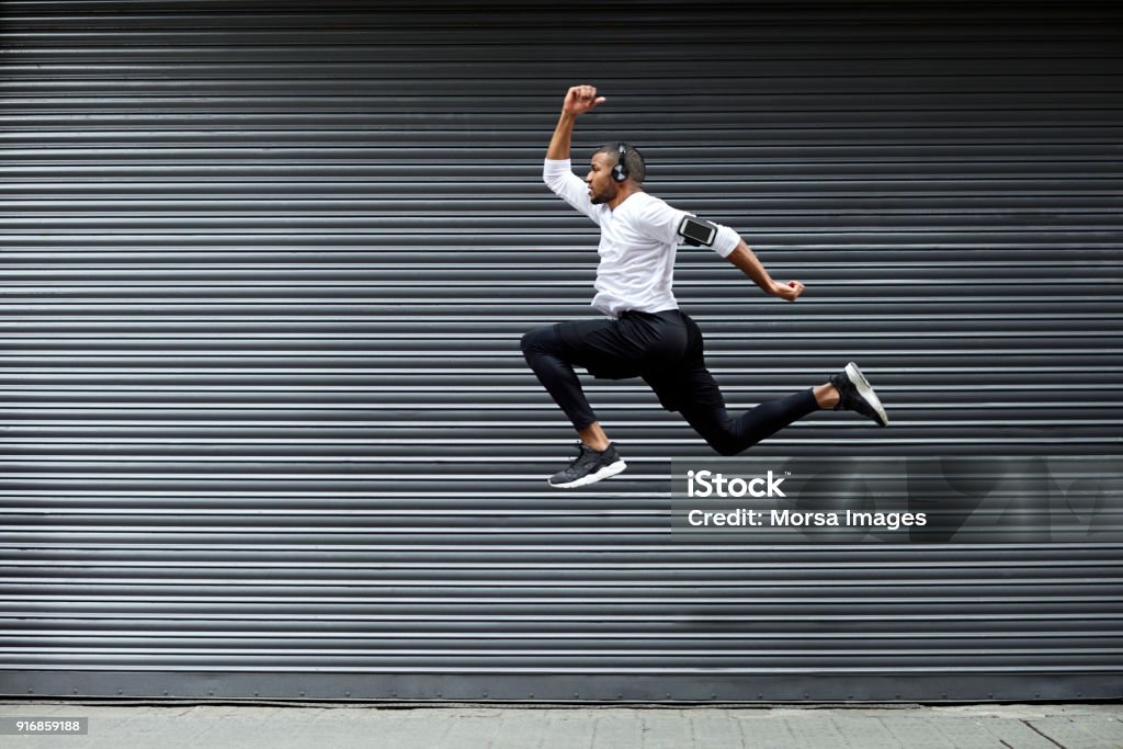 Sporty young man jumping against shutter Full length of determined young man jumping against shutter. Side view of fit male jogger is exercising in mid-air. He is wearing sports clothing. Jumping Stock Photo