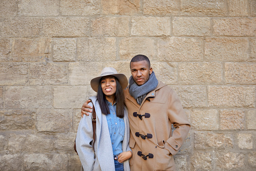 Portrait of couple standing against wall. Man and woman are in warm clothing. They are in city.