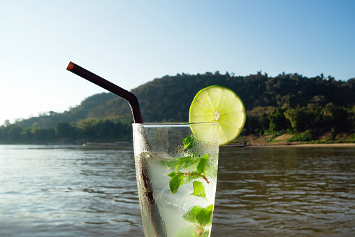 A glass of iced mojito with lime and leaves of mint on a background of river during of sunset. Luangprabang, Laos.