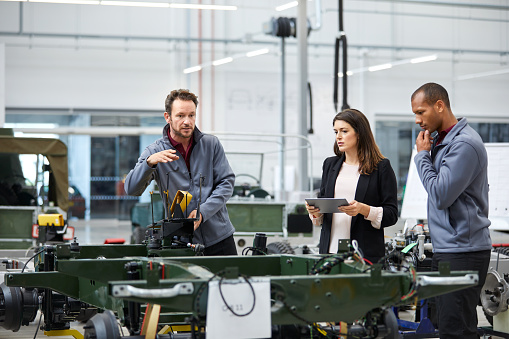 Automobile engineer showing incomplete car while discussing with colleagues. Multi-ethnic male and female professionals are standing at car production line. They are in automotive industry.