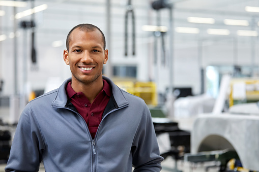 Portrait of maintenance engineer smiling in automobile industry. Male technologist is standing in showroom. Confident professional is wearing jacket in factory.