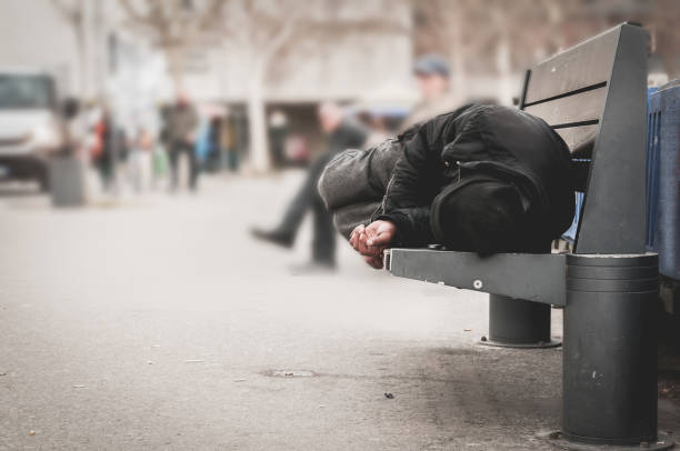 Poor homeless man or refugee sleeping on the wooden bench on the urban street in the city, social documentary concept, selective focus stock photo