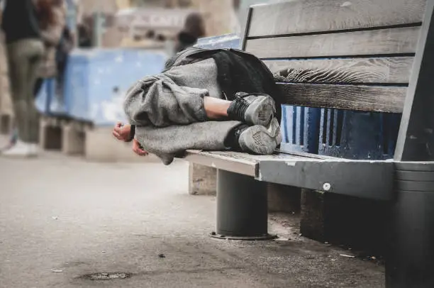 Poor homeless man or refugee sleeping on the wooden bench on the urban street in the city, social documentary concept, selective focus