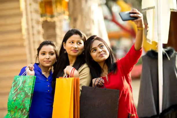 Indian women holding shopping bags and taking selfie using mobile camera at market