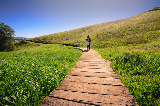 A mother and daughter going for a hike through a meadow. They're walking along a wooden foot path, surrounded by lush green grass on either side. The location is set in the hills of Orange County, CA after a recent Winter rainfall.