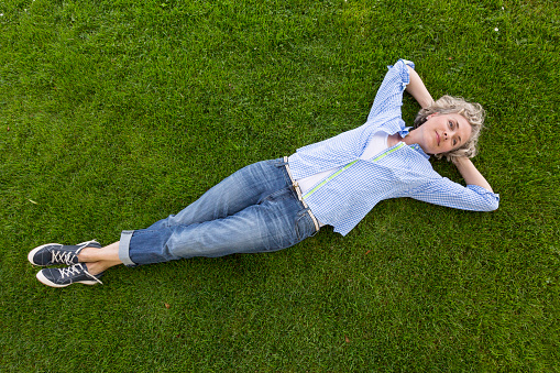 Middle-aged woman in casual weekend clothing relaxing on a grass lawn in a yard or park. She is smiling with a happy, contented expression and looks like she is daydreaming.