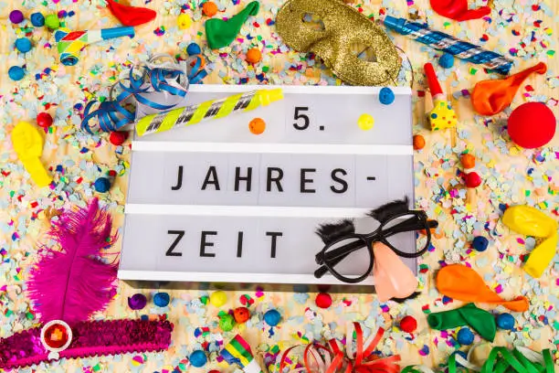 Lightbox with letters - 5. Jahreszeit means HAPPY CARNEVAL - on colorful festive party decoration with steamers, confetti and ballons.