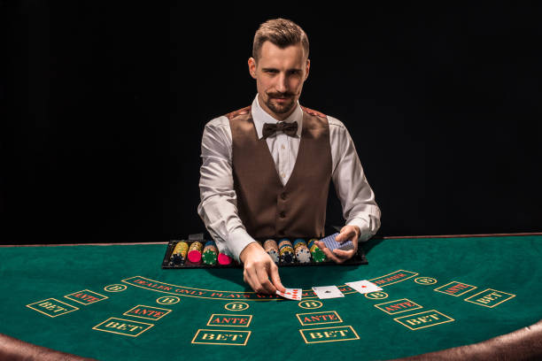 il ritratto di un croupier tiene le carte da gioco, le fiches da gioco sul tavolo. sfondo nero - croupier foto e immagini stock