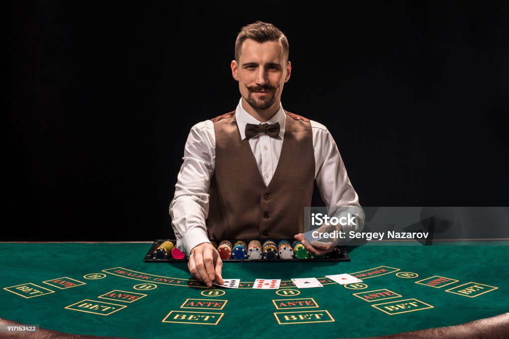 Portrait of a croupier is holding playing cards, gambling chips on table. Black background Portrait of a croupier is holding playing cards, gambling chips on table. Black background. A young male croupier in a shirt, waistcoat and bow tie is waiting for you at the blackjack table Casino Worker Stock Photo