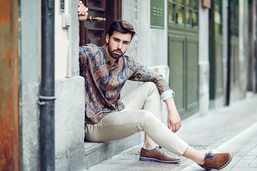 Young bearded man, model of fashion, sitting in an urban step wearing casual clothes. Guy with beard and modern hairstyle looking away in the street.