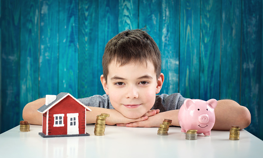 eight years old child sitting at the table with money and a piggybank. Happy boy with euro coins and house model