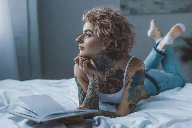 Photo of thoughtful tattooed girl lying on bed with book