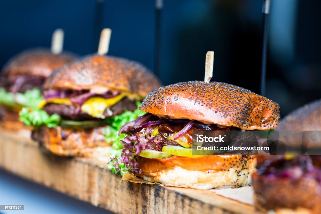 Fresh flame grilled burgers displayed in a row at food market Close up image of a selection of freshly flame grilled burgers in a row on a wooden counter at Borough Market, one of the oldest and most famous food markets in the world. Each of the burgers has its own label, on which is written the contents of the burger. The burgers are sandwiched between glazed buns, and presented on beds of fresh green lettuce and stuffed with melted cheese and red onion. Horizontal colour image with copy space. Burger Stock Photo