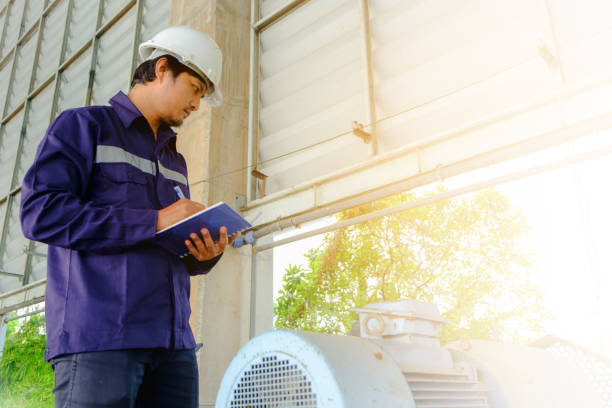 asian engineer or technician in blue collar safety uniform and white security helmet checking motor gear machinery equipment and writing data on notebook with sun light effect for industrial concept - ball bearing engineer machine part gear imagens e fotografias de stock