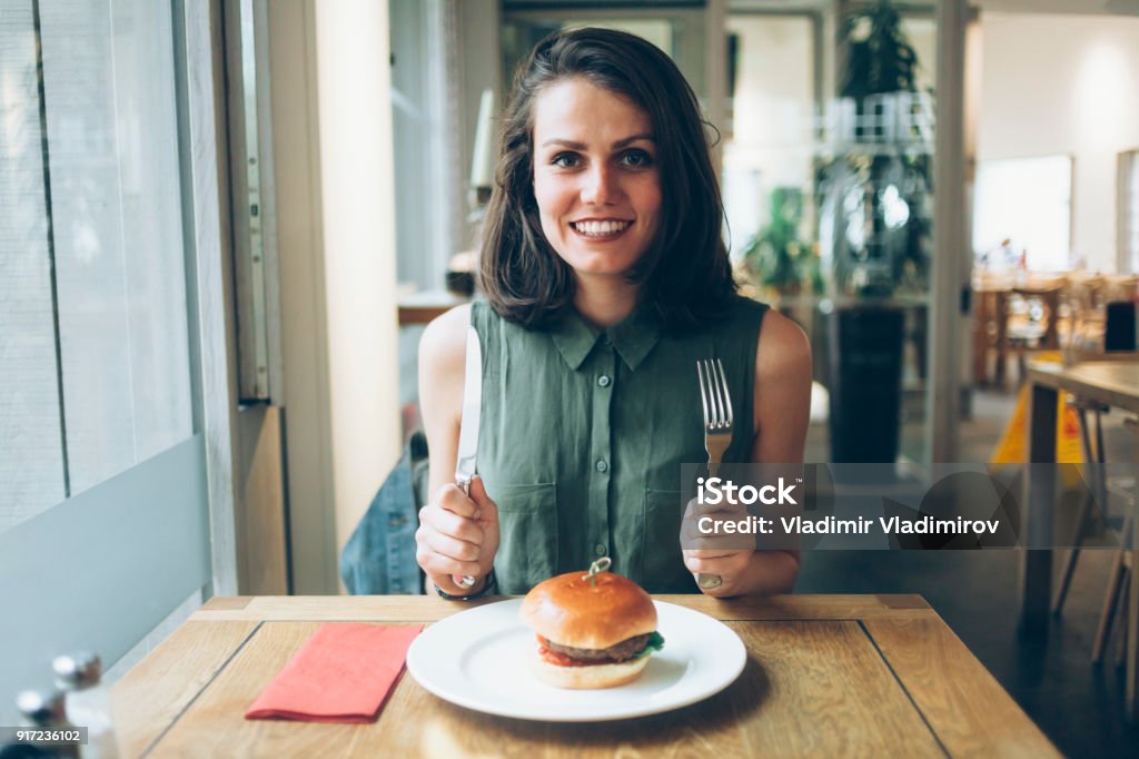 Elegant woman eating burger in restaurant Hungry young woman having lunch in restaurant, holding knife and fork. Looking at camera. Eating Stock Photo