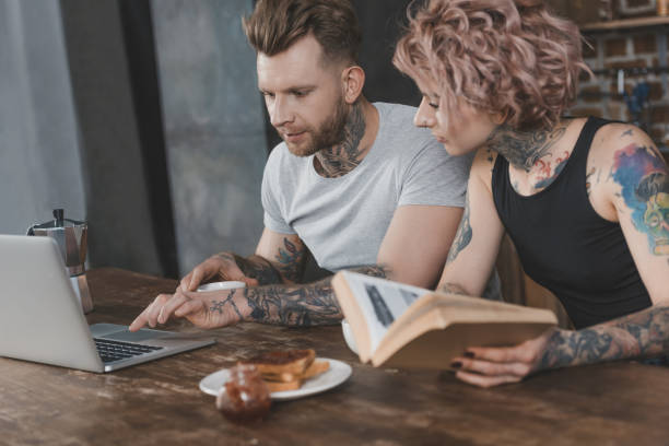 tattooed couple using laptop and reading book during breakfast in kitchen