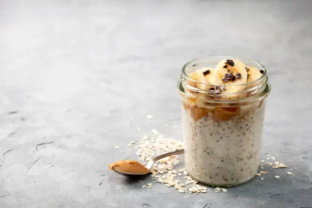 healthy diet breakfast. overnight oatmeal with chia seeds, bananas, peanut butter, honey, chocolate sprinkling in a glass jar on a gray concrete background