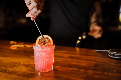 Bartender holding tongs in his hands adorns pink cocktail with an grapefruit slice. Barman at work, preparing cocktails