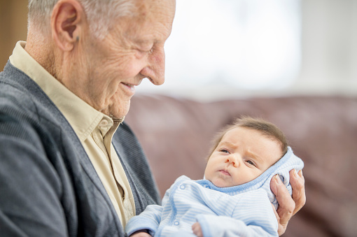 A Caucasian grandfather and his newborn grandson are indoors in a living room. The grandfather is smiling and holding the baby in his arms white sitting on the couch.