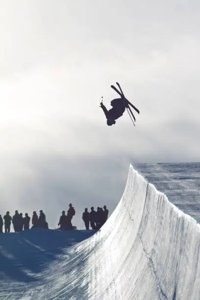A male alpine skier does a backflip off a half pipe jump while other skiers look on from above.