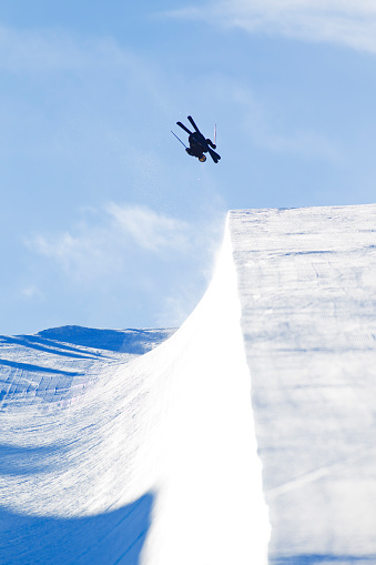 A male alpine skier does a backflip off a half pipe jump on a sunny day in the wintertime.