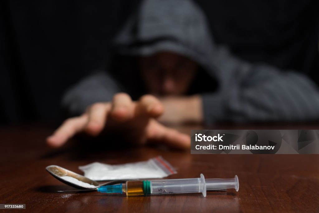 Addict at the table pulls his hand to the syringe with the dose Addict at the table pulls his hand to the syringe with the dose. Copy paste Drug Abuse Stock Photo