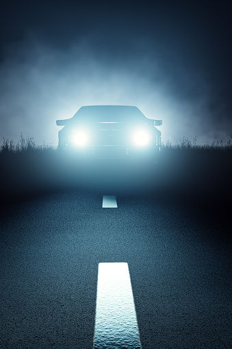 Car lights seen from the front on a dark eerie misty night and approaching on a striped asphalt road (3D render)