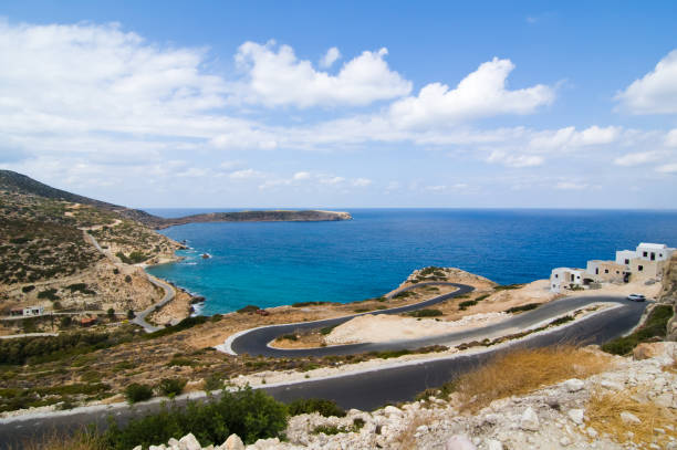 Landscape panorama of a winding road down a hill leading to the sea. Deep azure sea and cloudy sky in the background. Small pueblo on the side of the photo.