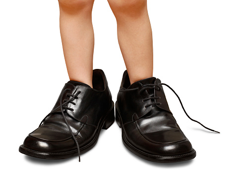 A preschooler tries on adult sized shoes while pretending to be a grownup.  The mens dress shoes are shown on the wrong feet with laces untied. File carefully cleaned up and isolated on a white background.