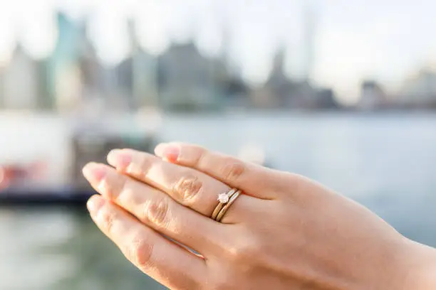 Young woman's hand with diamond engagement ring solitaire, gold wedding band outside outdoors in NYC New York City Brooklyn Bridge Park by east river, cityscape, skyline bokeh