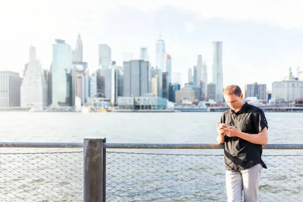Young man outside outdoors in NYC New York City Brooklyn Bridge Park by east river, railing, looking at view of cityscape skyline, texting on social media phone