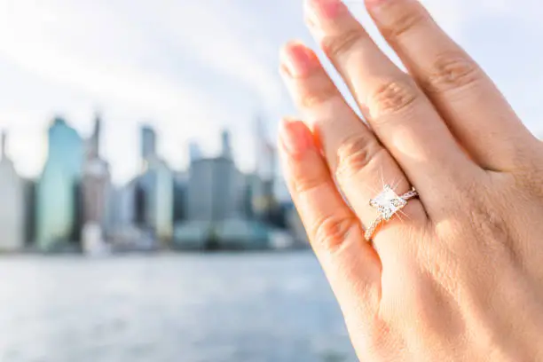 Young woman's hand with diamond engagement ring princess cut, gold outside outdoors in NYC New York City Brooklyn Bridge Park by east river, cityscape, skyline bokeh